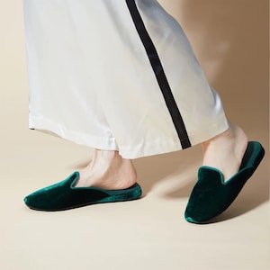 May include: A pair of dark green velvet slippers with a black trim. The slippers are worn by a person wearing white pants with a black stripe.