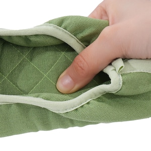 May include: A sage green quilted fabric slipper featuring a soft, textured surface with a diamond-stitched pattern and a light green fabric trim along the opening. The material appears flexible and padded, showing a close-up view of the textile.