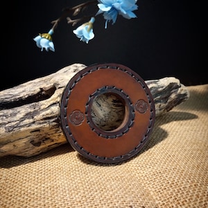 May include: A brown leather ring with a decorative stitch pattern and two floral designs. The ring is on a piece of driftwood.