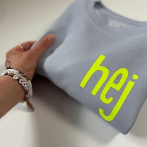 Puede incluir: Una sudadera gris claro con la palabra "hej" impresa en amarillo brillante.
