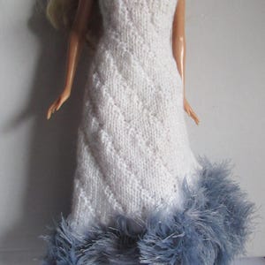 Peut inclure: Une robe blanche en tricot sans bretelles avec une bordure en plumes bleues. La robe a un motif en losanges et est faite pour une poupée.