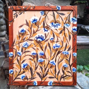 May include: A rectangular wooden frame containing a painting on a light peach background. The artwork features a repeating pattern of dark brown leafy stems and numerous small, rounded white flowers with vibrant blue centers and white dots.