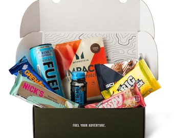 May include: An open cardboard box filled with various energy bars, protein wafers, and a blue raspberry energy drink. The box also contains a small bottle of workout supplement. The text "FUEL YOUR ADVENTURE" is printed on the box.