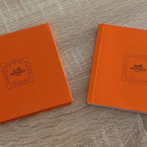 May include: Two square books with bright orange covers. Each cover features the text "CARRÉ" printed in a square formation around the central "HERMÈS PARIS" logo. The books have a textured finish and are positioned side by side on a flat surface.