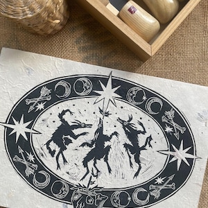 May include: Black and white linocut print of three stylized figures dancing around a star. The figures are surrounded by a circle of moons, stars, bones, and arrows.