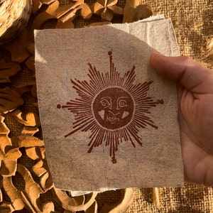 May include: A square, beige fabric featuring a brown sun design. The sun has a face and radiating lines. The fabric is held up, revealing a glimpse of a wooden carved design and a woven background.