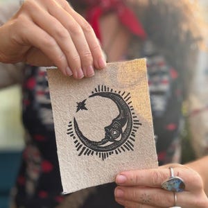 May include: A beige card with a black crescent moon design, a star, and sun rays. The card is held up by a person. The image has a vintage, bohemian aesthetic.
