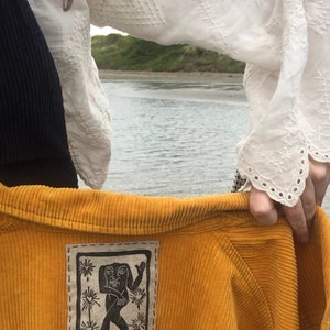 May include: A mustard yellow corduroy jacket with a black and white embroidered patch featuring a stylized human figure. The patch is sewn onto the back of the jacket.