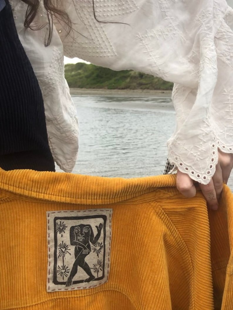 May include: A mustard yellow corduroy jacket with a black and white embroidered patch featuring a stylized human figure. The patch is sewn onto the back of the jacket.