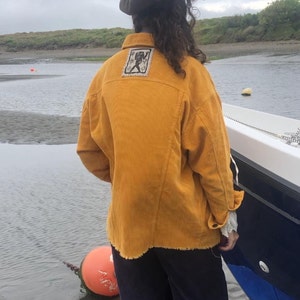 May include: A mustard yellow corduroy shirt with a frayed hem and a small embroidered patch on the back. The person is wearing black pants and a brown corduroy cap.