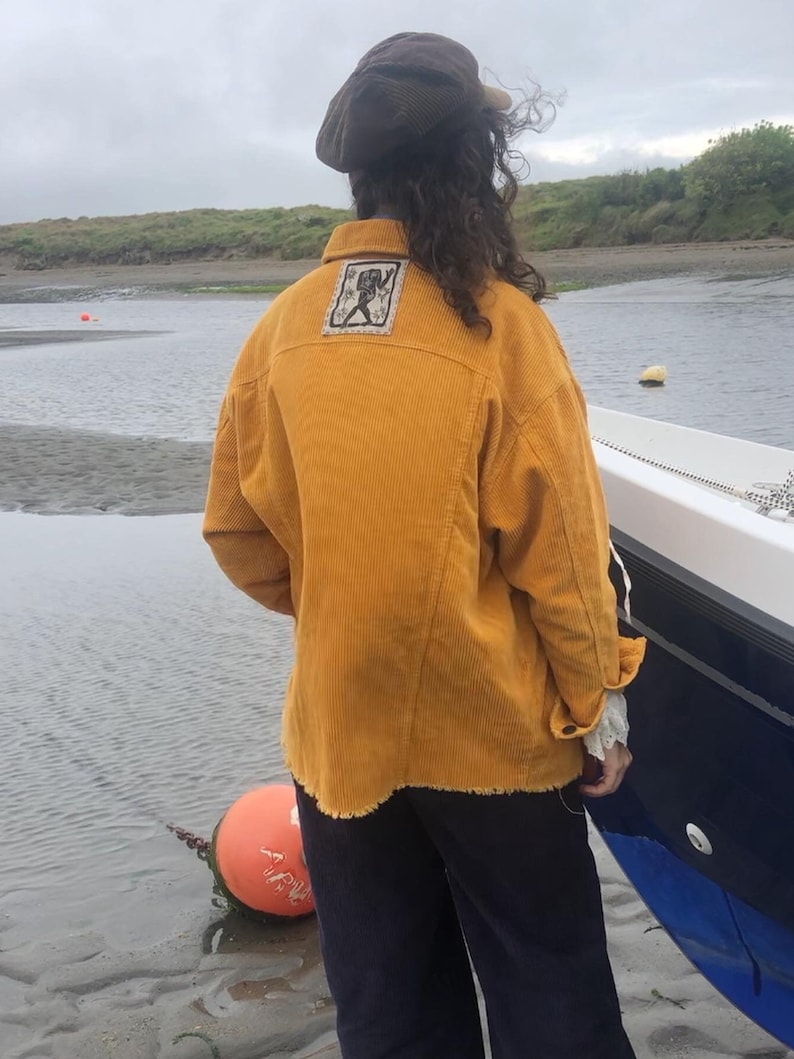 May include: A mustard yellow corduroy shirt with a frayed hem and a small embroidered patch on the back. The person is wearing black pants and a brown corduroy cap.