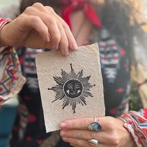 May include: A beige rectangular card featuring a black sun design with radiating lines and a smiling face. The card is held up by a person. The person is wearing rings and a patterned top.
