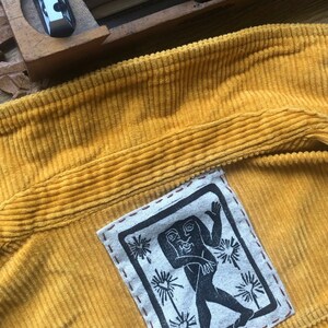 May include: A yellow corduroy tote bag with a black and white embroidered patch featuring a stylized figure with a raised hand and a geometric design.