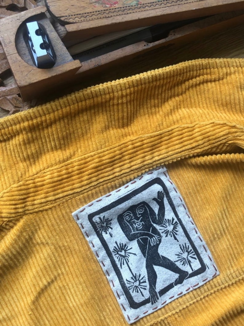 May include: A yellow corduroy tote bag with a black and white embroidered patch featuring a stylized figure with a raised hand and a geometric design.