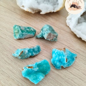 Könnte beinhalten: Fünf Stücke leuchtend türkisfarbener roher Edelsteine mit kristalliner Struktur. Die Edelsteine werden auf einer hellen Holzoberfläche mit einer weißen Geode im Hintergrund präsentiert. Ein Logo mit dem Text "Lumiere de Gemmes" befindet sich oben rechts.