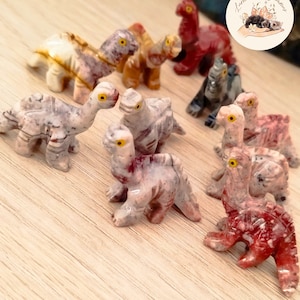 Puede incluir: Una colección de pequeñas figuras de dinosaurios talladas en varios colores, incluyendo rojo, marrón y gris. Cada figura tiene ojos amarillos y se exhibe sobre una superficie de madera clara. Los dinosaurios están en diferentes poses.