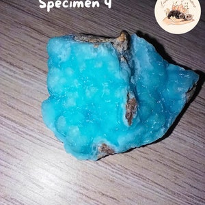May include: A vibrant, turquoise mineral specimen with a rough, textured surface. The stone is labeled "Sp&eacute;cimen 4" in white script. A circular logo with the text "Lumi&egrave;re de Gemmes" and a small illustration is in the upper right corner.