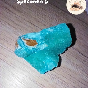 May include: A vibrant turquoise mineral specimen with a rough, textured surface. The stone has patches of brown and is labeled "Sp&eacute;cimen 5". The image also includes a logo with the text "Lumi&egrave;re de Gemmes".