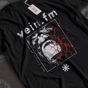 Könnte beinhalten: Ein schwarzes T-Shirt mit einer weißen Grafik eines schreienden Gesichts in einem quadratischen Rahmen. Der Text "vein.fm" steht über dem Rahmen und "pain. pressure. permanent." ist an der Seite aufgedruckt. Ein rotes Adernmuster überlagert das Gesicht.