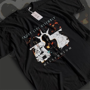Może przedstawiać: Czarny t-shirt z krótkim rękawem z nadrukiem graficznym zawierającym tekst FRONT LINE ASSEMBLY oraz MILLENNIUM. Wzór zawiera biały abstrakcyjny kształt z pomarańczowymi i niebieskimi akcentami. Przy kołnierzu znajduje się biała metka w rozmiarze L.