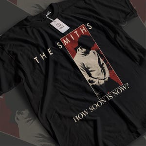Può includere: Una t-shirt nera a maniche corte con una stampa grafica di una persona con un maglione a collo alto all'interno di una cornice rettangolare rossa. Il testo THE SMITHS appare sopra la grafica e HOW SOON IS NOW? è stampato sotto. È presente un'etichetta taglia large.