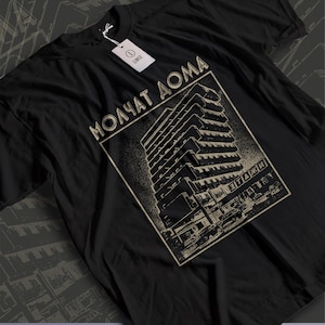 May include: A black short-sleeve t-shirt featuring a graphic print of a brutalist building with the Cyrillic text "МОЛЧАТ ДОМА" and "ЭТАЖИ" in a beige color. The shirt includes a white paper tag attached to the collar with the letter L printed on it.
