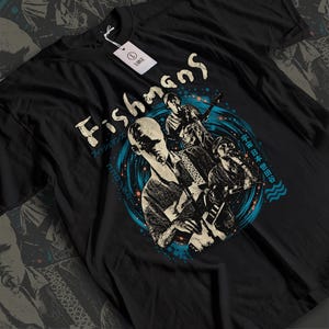 以下が含まれることがあります： 3人のミュージシャンが描かれたグラフィックプリントの黒い半袖Tシャツ。中央のイラストの上部には「Fishmans」の文字がスタイリッシュなフォントで配置され、周囲には日本語の文字と青い円形のアクセントがデザインされています。