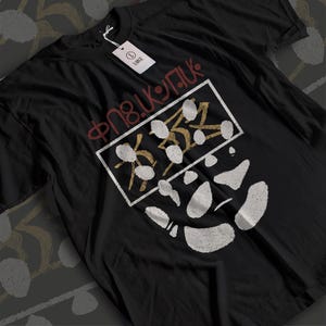 May include: A black short-sleeve t-shirt featuring a white rectangular frame containing gold and white abstract shapes. Above the frame is red text that reads "ΦΠ8.ΚΨΠΚ". A white tag attached to the collar displays the letter "L" and the word "LARGE".