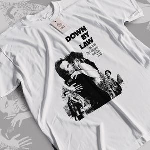 Könnte beinhalten: Ein weißes kurzärmeliges T-Shirt mit einem schwarz-weißen Grafikdruck von Szenen aus dem Film Down by Law. Der Text lautet Down by Law und sous le coup de la loi. Ein kleines rechteckiges Etikett mit dem Buchstaben L ist am Kragen befestigt.