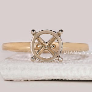 Puede incluir: Un anillo de oro con un engaste plateado diseñado para sostener una piedra preciosa. El anillo tiene una banda simple y un engaste detallado con cuatro garras. El anillo se muestra sobre una superficie blanca.