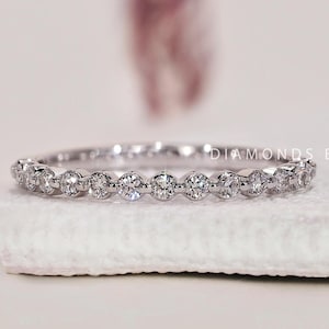 May include: A delicate eternity band with a row of small round diamonds set in a white gold band. DIAMONDS BY UK is printed on the white surface behind the ring.