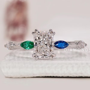 Könnte beinhalten: Ein silberner Ring mit einem großen, rechteckigen, klaren Edelstein als Mittelstück, flankiert von einem grünen und einem blauen Edelstein. Kleine, klare Edelsteine sind entlang des Bandes gesetzt. Der Text "DIAMONDS BY UK" ist sichtbar.