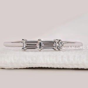 Puede incluir: Un delicado anillo de plata con tres piedras preciosas claras de corte rectangular y una pequeña piedra redonda. El anillo está colocado sobre una superficie blanca, con el texto "DIAMONDS BY UK" visible en el fondo.