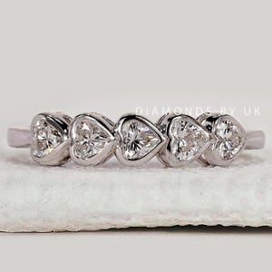 Peut inclure: Une bague en argent ornée de cinq diamants en forme de cœur. Les diamants sont disposés en ligne, créant un motif continu. La bague est présentée sur une surface blanche. Le nom de la marque "DIAMONDS BY UK" est visible.