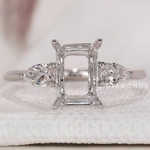May include: A silver ring setting with an emerald-cut center and pear-shaped side stones. The ring is set on a white surface. The ring is designed to hold gemstones. The text "DIAMONDS BY UK" is visible in the background.