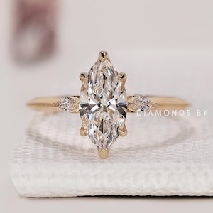 May include: A gold engagement ring featuring a large, marquise-cut diamond as the centerpiece. The ring has smaller diamonds on either side of the main stone. The band is a simple, polished gold. The ring is set on a white surface.