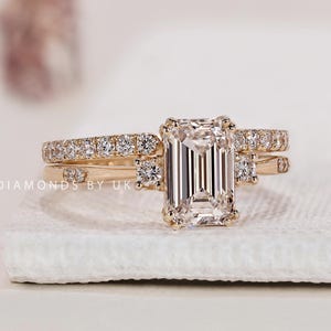 Op de afbeelding: Een gouden ringenset met een centrale diamant in emerald-slijpvorm, geflankeerd door kleinere ronde diamanten op de ringband. De band is bezet met pavé-diamanten en de tekst DIAMONDS BY UK is zichtbaar aan de zijkant van de zetting.