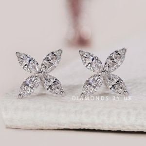 May include: A pair of silver-toned stud earrings, each featuring four marquise-cut clear stones arranged in a flower-like cluster. The text DIAMONDS BY UK is visible in the lower right corner, and a logo is present in the upper left corner.