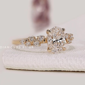 Op de afbeelding: Een gouden verlovingsring met een grote ovaal geslepen diamant en kleinere diamanten op de band. De ring ligt op een wit oppervlak. De merknaam "DIAMONDS BY UK" is zichtbaar.