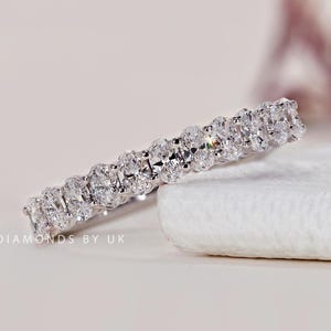 Pode incluir: Um anel de eternidade de diamantes com uma fileira contínua de diamantes de corte oval. O anel, cravejado em metal branco, é exibido sobre uma superfície branca. Os diamantes brilham, refletindo a luz.