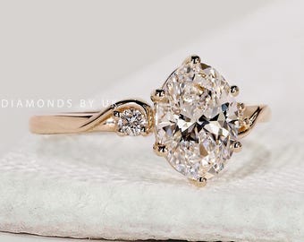 May include: A gold engagement ring with a large, oval-cut diamond as the centerpiece. The band features a delicate, swirling design with a smaller diamond accent. The ring is set on a white surface.