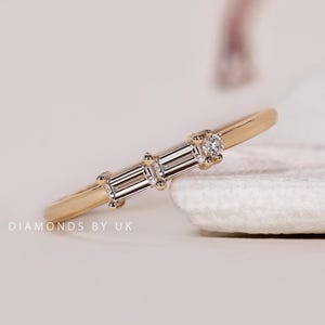 Könnte beinhalten: Ein goldener Ring mit drei Diamanten. Der Ring hat einen zentralen Diamanten im Baguetteschliff, flankiert von zwei kleineren Diamanten. Der Ring liegt auf einer weißen Oberfläche. Der Text "DIAMONDS BY UK" ist sichtbar.