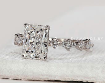 May include: A radiant-cut diamond engagement ring. The ring features a large, rectangular-shaped center stone and smaller accent diamonds along the band. The ring is set in a silver or white gold band, with the brand name "DIAMONDS BY UK" visible.