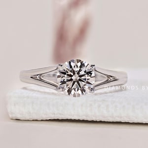 Op de afbeelding: Een close-up van een diamanten ring met een split-shank design. De ring heeft een grote, rond geslepen diamant gezet in een witte metalen band. De band splitst aan weerszijden van de diamant, wat een unieke uitstraling creëert. De ring wordt getoond op een witte ondergrond.