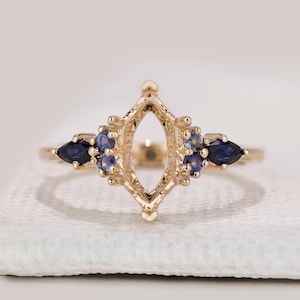 May include: A gold ring featuring a central marquise-shaped empty setting flanked by two dark blue pear-cut stones and clusters of smaller round blue stones on each side. The metal band is polished gold, creating a symmetrical, decorative design.