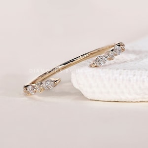 May include: A gold-colored ring with multiple clear gemstones. The ring has a unique open design, with gemstones set along the band. The ring is displayed on a white surface.