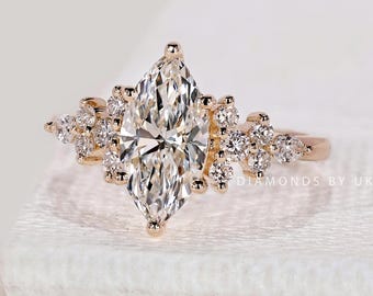 Könnte beinhalten: Ein Verlobungsring aus Roségold mit einem Diamanten im Marquiseschliff im Zentrum. Der Ring ist mit kleineren Diamanten verziert, die sich um den Hauptstein und entlang des Bandes gruppieren. Der Ring liegt auf einer weißen Oberfläche, mit dem Text "DIAMONDS BY UK" sichtbar.