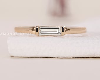 May include: A gold band ring with a rectangular, clear gemstone. The ring is set on a white surface. The words "DIAMONDS BY UK" are printed on the left side of the image.