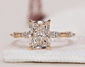 May include: A radiant-cut diamond engagement ring with a gold band. The ring features a large rectangular center stone and smaller accent stones on the band. The ring is set on a white surface.