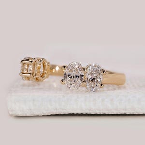 May include: A gold ring with three oval-cut diamonds and a smaller cluster of diamonds. The ring is set on a white surface, showcasing the sparkle of the gemstones. The band is a simple, polished gold.